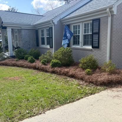 Front yard of a house with green shrubs, pine straw, and a flag on a sunny day.